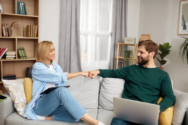 Couple Using Gadgets At Home. Married Couple Of Freelancers Bumping Hands While Work From Home. Guy And Girl In Casual Clothes With Gadgets Work In The Living Room At The Sofa
