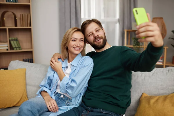 自宅でセルフィーを取るカップル 自宅でソファでリラックスしながら携帯電話を使用して幸せなカップル 現代のスマートフォンを使って興奮した人たちが一緒に笑う — ストック写真