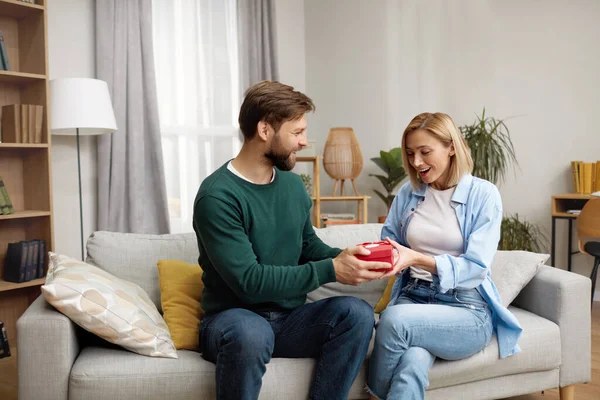 Happy Man Presenting Gift To Wife. Excited Young Man Presenting Good Unexpected Present To Wife At Home. Loving Boyfriend Making Romantic Surprise To Girlfriend