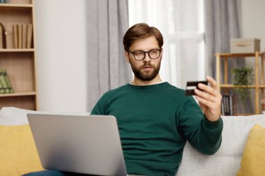 Ciddi Adam Tutma Kredi Kartı. İnternette sakallı adam ödeme yapıyor, dizüstü bilgisayar kullanıyor, plastik kredi kartı tutuyor, evdeki koltukta oturuyor. Genç Erkek Alışveriş, Güvenli İnternet Ödemesi 