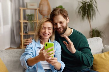 Evde selfie çeken bir çift. Mutlu çift evdeki koltukta dinlenirken cep telefonu kullanıyor. Heyecanlı İnsanlar Modern Akıllı Telefonu Birlikte Kullanarak Güler 