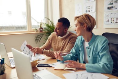 Smiling Colleagues Working At Office. Happy Business Couple Discussing Documents Using Computer In Modern Office. Woman Giving Advices To Man. Teamwork Concept