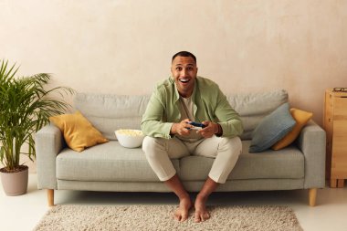 Overjoyed Man Playing Video Games. Multiracial Guy Sitting On Couch In Living Room Enjoying Playing Video Games. Enjoying Moment On Weekend Concept 