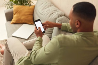 Man Using Smartphone At Home. Handsome Multiracial Guy Sitting On Sofa And Messaging In Cozy Living Room at Home. Male Person Doing Online Shopping, Browsing Internet and Checking Social Media 
