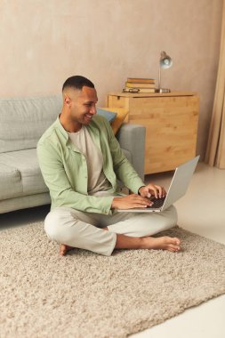 Smiling Man Using Laptop At Home. Young Guy Holding Computer, Sitting At Floor At Cozy Home. Multiracial Man Relaxing With Laptop Alone 