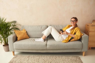 Woman Using Smartphone At Home. Blonde Girl Sitting On Sofa And Messaging In Cozy Living Room at Home. Female Person Doing Online Shopping, Browsing Internet and Checking Social Media 