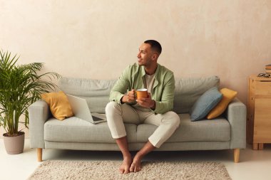 Happy Man Drinking Coffee. Relaxed Guy Sitting On Couch With Mug Of Tea In Cozy Living Room At Home. Domestic Lifestyle Concept 