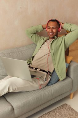 Headphones Man Listening Music. Cheerful Guy Using Laptop At Home. Smiling Millennial Man Sitting On Couch In Living Room And Using Pc Computer, Listening To Music In Wireless Headset 