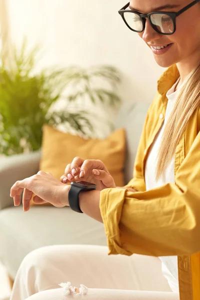 Smiling Woman Using Smart Watch. Caucasian Girl Touching Wrist Watch While Sitting Sofa In Living Room. Positive Person Trying New Technologies