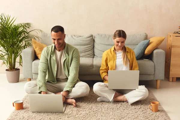 Couple Using Laptops At Home. Married Couple Of Freelancers Work From Home. Guy And Girl In Casual Clothes With Gadgets Work In The Living Room At The Floor