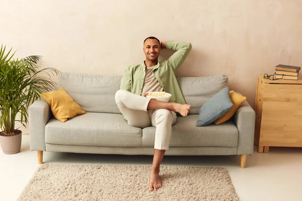 Happy Man Relaxing Sofa. Smiling Middle Eastern Guy Sitting On Comfortable Couch And Eating Pop Corn At Home In Living Room. Cheerful Man Relaxing On Sofa, Enjoying Weekend