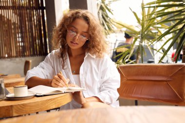 Freelancer At Cafe. Serious Woman In Glasses Writing Something in Notebook. Remote Job Or Education With Modern Digital Technologies For Comfortable Lifestyle In City