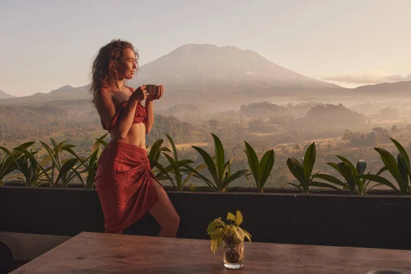 Woman enjoys coffee on yoga retreat at a luxurious resort with stunning views of Agung volcano and the beautiful sunrise in Bali during her travels.
