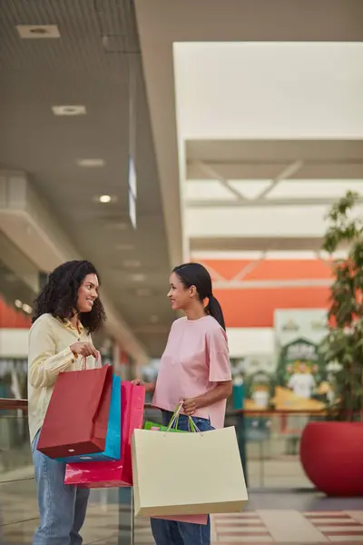 Two people walking, carrying shopping bags, speaking, and smiling. They are happy friends enjoying a discount sale at a mall store.