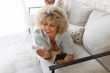 Neşeli kıvırcık saçlı genç bir kadın kanepeye uzanmış elinde akıllı bir telefon ve kartla evde rahatlatıcı bir anın tadını çıkarıyor..