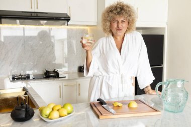 Beyaz bornozlu bir kadın, modern bir mutfakta elinde bir bardak su tutarken suratını ekşitiyor. Tezgahında taze limonlar var..