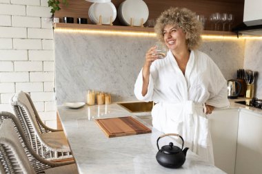 Beyaz bornozlu neşeli genç bir kadın modern bir mutfakta detoks suyunu yudumluyor ve evde sağlıklı bir sabah rutini için olumlu bir hava yaratıyor..