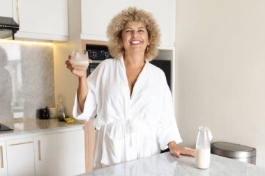 Bornozlu neşeli kıvırcık saçlı bir kadın modern mutfak ortamında bir bardak sütün tadını çıkarıyor, pozitif ve sağlıklı..