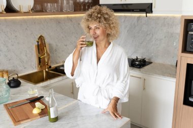 Beyaz cüppeli kıvırcık saçlı bir kadın şık bir mutfak ortamında sağlıklı yeşil bir meyve suyu yudumluyor, rahatlatıcı ve sağlıklı..