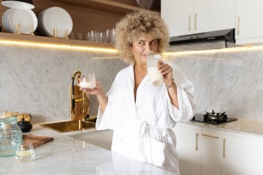 Bornozlu, kıvırcık saçlı, mutlu bir kadın. İyi ışıklandırılmış, zarif bir mutfak ortamında süt içiyor. Sağlıklı bir yaşam tarzı yayıyor..