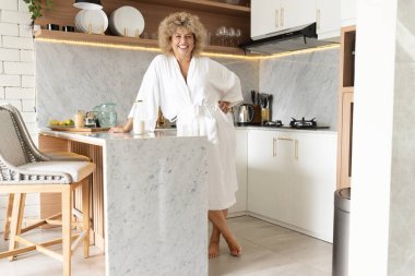 Beyaz cüppeli neşeli bir kadın modern bir mutfakta duruyor, konforu somutlaştırıyor ve evde rahat bir yaşam sürüyor, çağdaş iç tasarım unsurlarıyla birlikte..