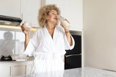 Rahat, bornoz giymiş olgun bir kadın şık, şık bir mutfak ortamında kokulu bir mum kokluyor..