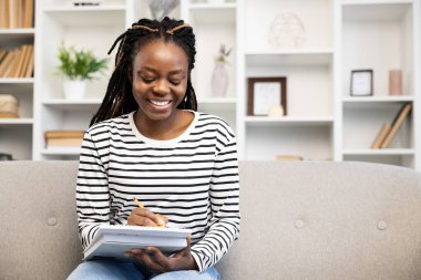 Evdeki kanepede oturan neşeli Afro-Amerikan kadın, hobileriyle meşgul. Konforlu, dekore edilmiş bir yaşam alanıyla çevrili bir not defterine mutluluk ve memnuniyet dolu bir yazı yazar..