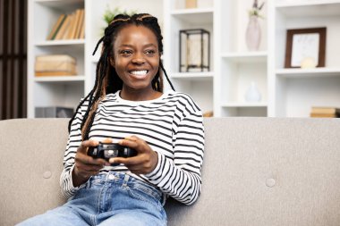 Saçları örgülü neşeli Afro-Amerikalı kadın kanepesinde rahat rahat oturup, rahat bir oturma odasında keyif ve rahatlık sunuyor..