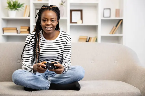 アフロ系アメリカ人女性がソファーに足を踏み入れ 居心地の良いリビングルームでビデオゲームのコントローラーと深く関わり モダンな趣味とレジャータイムを披露します — ストック写真