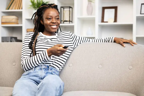 Happy African American woman relaxing at home, comfortably sitting on sofa and holding a remote control, with cozy home interior in the background.