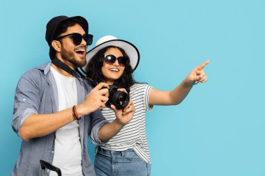 Kameralı neşeli turist çifti mavi bir arka plana bakıp gülüyorlar, güneş gözlüğü ve şapka takıyorlar..