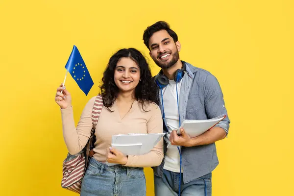 在黄色背景下 快乐的年轻学生举着印有欧盟旗帜的学习用品 — 图库照片