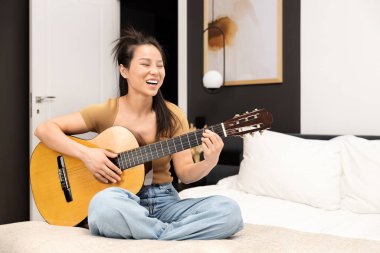 Yatak odasında akustik gitar çalan neşeli Asyalı kadın. Günlük ev eğlencesi, müzik hobisi, boş zaman konsepti. Mutlu, rahatlatıcı, müzikal enstrüman, öğrenme, uygulama.