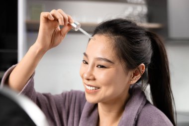 Gülümseyen Asyalı kadın evde cilt bakımı için serum kullanıyor. İçerik ve Tazelik, Düşüren Şişe ile Günlük Güzellik Muamelesi. Kişisel Bakım ve Refah Konsepti.