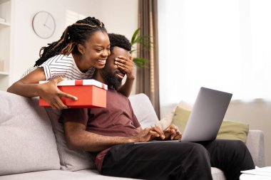 Sürpriz Hediye Zamanı: Kutulu Erkek Sunan Kadın, Neşeli ve Sürprizin İfadesi, Evde Birlikte Eğlenen Çift, Laptop Yaşam Tarzı, Aşk ve İlişki Konsepti, Neşeli Afrikalı Amerikalı Çift