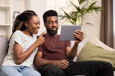Çift evde dijital tablet teknolojisinin tadını çıkartıyor. Rahatlamış Adam Tablet 'i tutarken Gülümseyen Genç Kadın ekranı işaret ediyor. Ev İçi Eğlence, Aşk İlişkileri ve Modern Yaşam Tarzı Konsepti. 