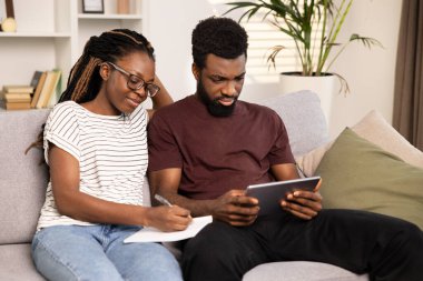 Çift birlikte plan yapıyor, Afro-Amerikan erkek ve kadın not yazıyor ve evde tablet kullanıyor. Takım çalışması, işbirliği, ev bütçesi planlama kavramı. Çeşitli, Mutlu, Teknoloji