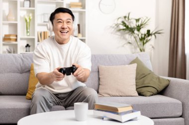 Mutlu Asyalı adam koltukta video oyunu oynuyor, evde boş zamanın tadını çıkarıyor, ev eğlencesi kavramı, kulaklıkla gülümseyen oyuncu ve joystick 'le eğleniyor.