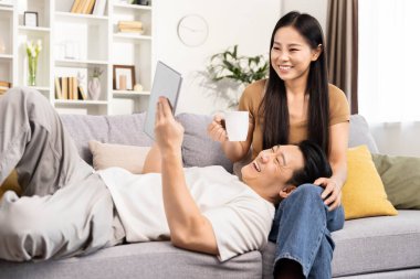 Memnuniyet duyan Asyalı çift evde dinleniyor, adam tabletle kanepeye uzanıyor, kadın kahve fincanı tutuyor, haftasonu teknolojiyle eğleniyor..