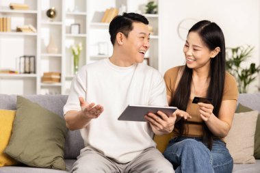 Mutlu Asyalı çift evdeki kanepede tablet ve kredi kartıyla online alışveriş yapıyor, teknolojinin keyfini çıkarıyorlar.