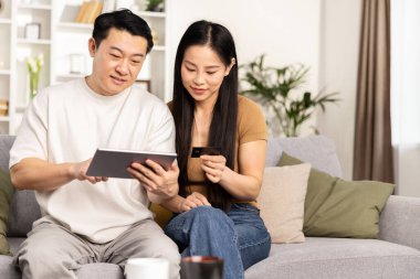 Kanepede oturmuş dijital tablet ve kredi kartıyla online alışveriş yapan Asyalı çift..