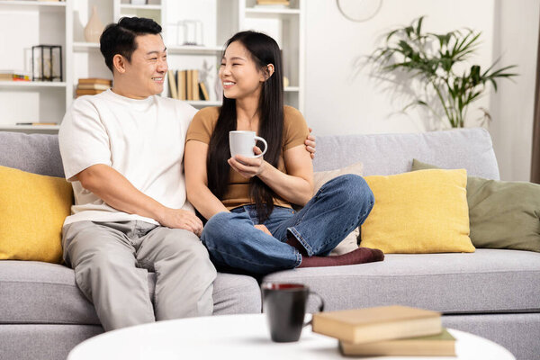 Счастливая азиатская пара расслабляясь вместе дома, участвуя в интимной беседе на удобном диване, показывая привязанность и радость, современная гостиная