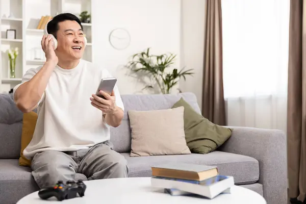 ハッピーマンは 家庭で音楽を楽しんでいます アジアの大人の笑顔現代リビングルームでスマートフォンやヘッドフォンを使用して カジュアル服 快適さ レジャー ポジティブバイブ ホームエンターテイメントコンセプト ストック画像