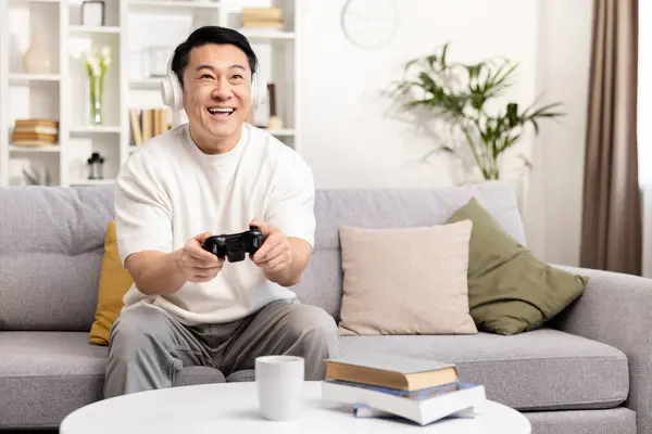 ハッピーアジアの男は カウチでビデオゲームをプレイ 楽しむレジャータイムインドア 楽しいホームエンターテイメントコンセプト ヘッドフォンとジョイスティックと笑顔のゲーマー ロイヤリティフリーのストック画像