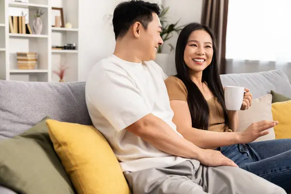 居心地の良いリビングルームで温かいコーヒーを楽しみながら ソファーに座っているアジアのカップルを微笑みます ストックフォト