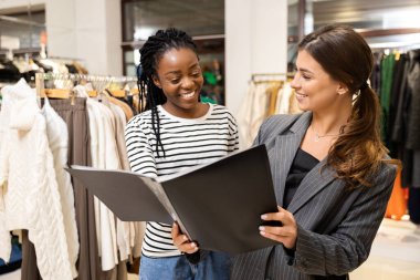 Butikte Danışman Alışverişi yapan, Kıyafetlerin Arasında Gülümseme Paylaşan Kadın. Mağazadaki Kadınlar Moda Kataloğunu Tartışıyor