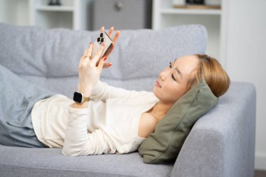 Genç bir kadın akıllı telefonuyla koltukta dinleniyor, evde boş zamanlarının, rahatlığın ve teknolojinin tadını çıkarıyor.