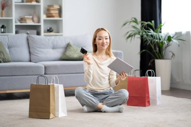 Çevrimiçi alışveriş yapan, tabletle yerde oturan, kredi kartı kullanan ve şık oturma odasında renkli alışveriş torbaları taşıyan genç bir kadın..