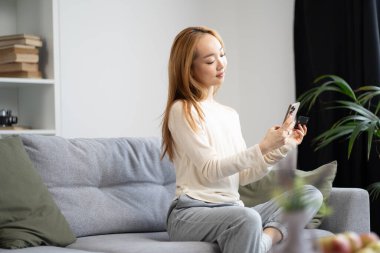 Akıllı telefon ve kredi kartı kullanarak koltukta dinlenen genç bir kadın, modern iç mimari. Sıradan Yaşam Tarzı, Dijital Bağlantı, Konforlu Yaşam Konsepti. Teknoloji ve Ev Konforu İçin Moda Görünümü.