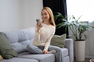 Genç bir kadın, modern oturma odasında akıllı telefonuyla koltukta keyif çatıyor, bitkiler ve doğal ışıkla çevrili, Neşe ve Bağlılık sergiliyor,.